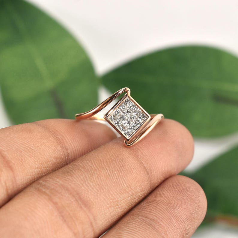Puede incluir: Un anillo de oro rosa con un cuadrado de peque&ntilde;as piedras brillantes. La banda del anillo tiene un dise&ntilde;o curvo, creando un aspecto moderno y elegante. El anillo se muestra sobre un fondo verde borroso.