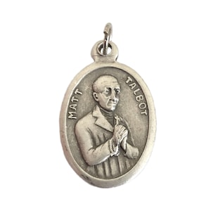 May include: Oval silver-toned pendant featuring a detailed relief of a man with the text "MATT TALBOT" around the edge. The man is depicted with clasped hands and a cross. The pendant has a small loop at the top for a chain.