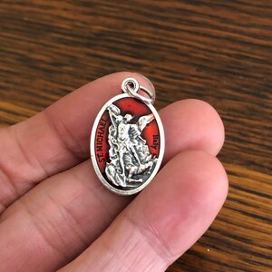 May include: An oval-shaped silver-toned pendant featuring a raised relief of Saint Michael defeating a demon against a vibrant red enamel background. The text ST. MICHAEL is printed on the left side and ITALY is printed on the right side.