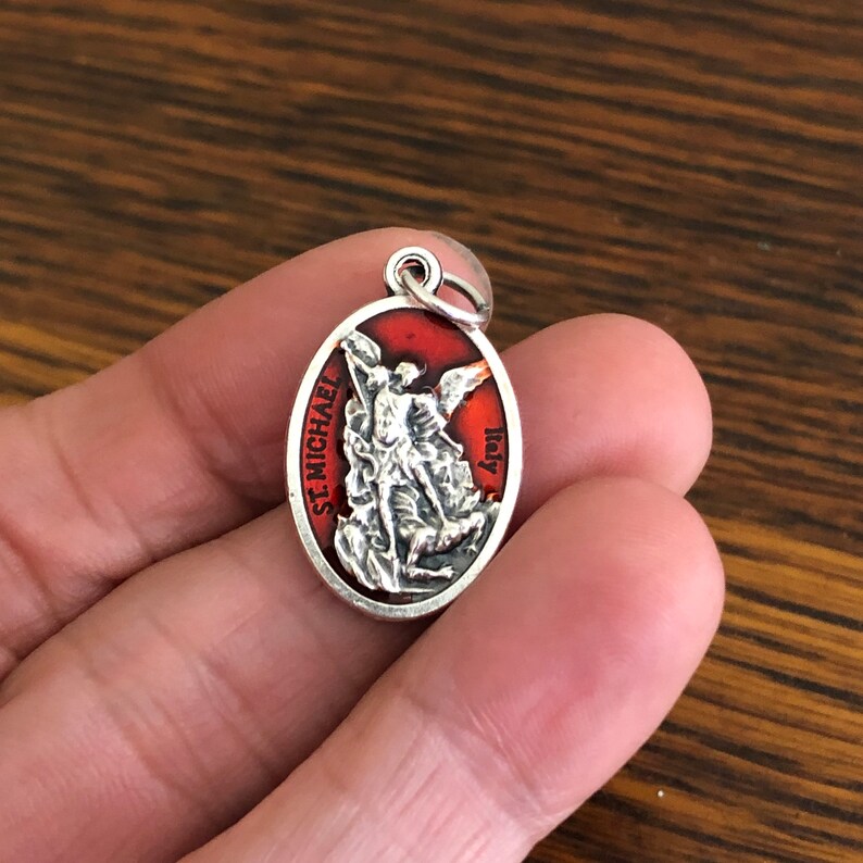 May include: An oval-shaped silver-toned pendant featuring a raised relief of Saint Michael defeating a demon against a vibrant red enamel background. The text ST. MICHAEL is printed on the left side and ITALY is printed on the right side.