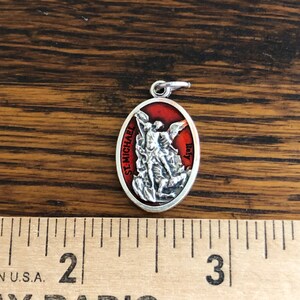 May include: An oval silver-toned pendant featuring a raised relief of Saint Michael defeating a demon against a red background. The text 'ST. MICHAEL' and 'Italy' is visible on the front. A ruler shows the pendant is approximately 1 inch tall.