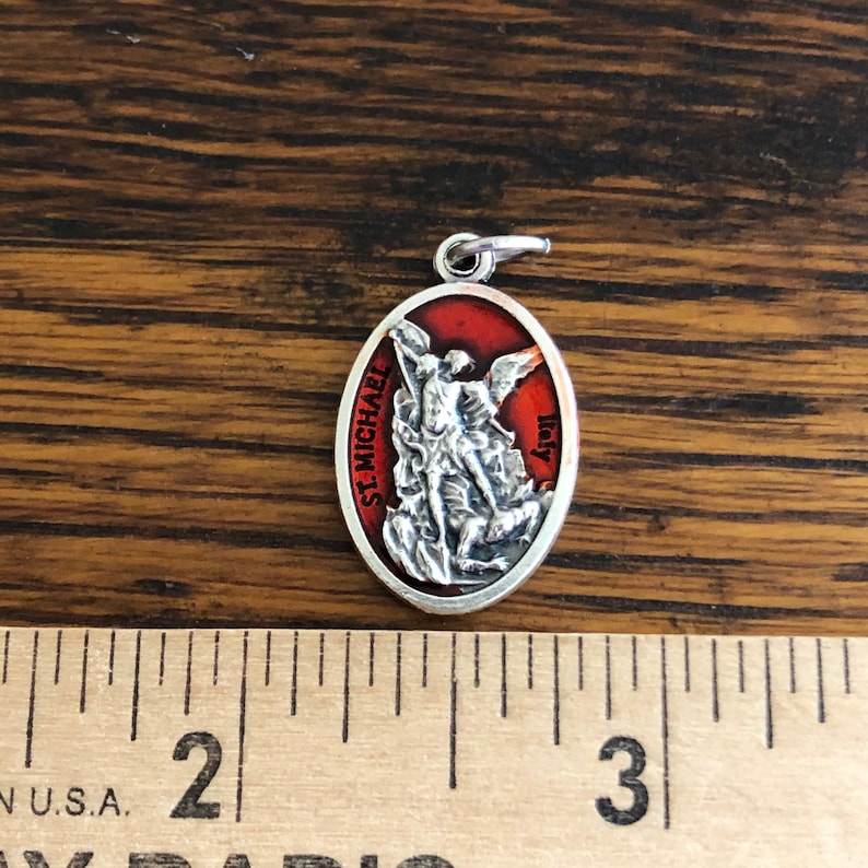 May include: An oval silver-toned pendant featuring a raised relief of Saint Michael defeating a demon against a red background. The text 'ST. MICHAEL' and 'Italy' is visible on the front. A ruler shows the pendant is approximately 1 inch tall.
