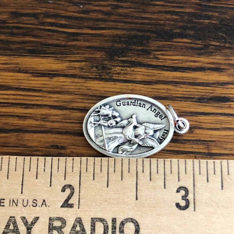 May include: An oval silver-toned metal charm featuring a relief of a guardian angel watching over two children. The text "Guardian Angel" and "Italy" are embossed on the surface. A metal jump ring is attached. A ruler shows the charm is approximately 1 inch wide.