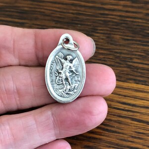 Silver color oval pendant featuring a depiction of Saint Michael the Archangel trampling the devil. The pendant has the words "St. Michael" inscribed on the side.