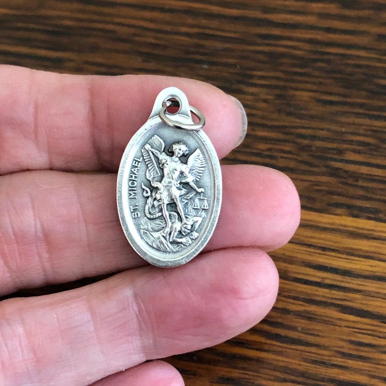 Silver color oval pendant featuring a depiction of Saint Michael the Archangel trampling the devil. The pendant has the words "St. Michael" inscribed on the side.