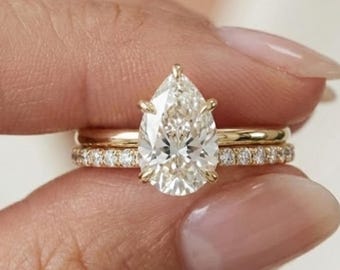 Puede incluir: Un conjunto de anillos de compromiso de oro con un diamante en forma de pera y una banda de diamantes. Los anillos se muestran, destacando el brillo de las piedras preciosas. El diseño es clásico y elegante.