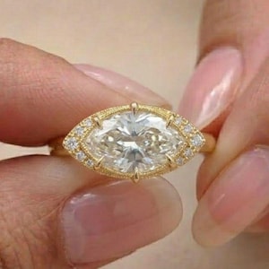 Puede incluir: Un anillo de oro con un diamante central de talla marquesa. El anillo está rodeado de diamantes de acento más pequeños y tiene una banda detallada y texturizada. El anillo se sostiene entre dos dedos.