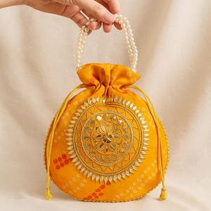 Op de afbeelding: Een gele trekkoordtas met een parelhandvat. De tas heeft een cirkelvormig, geborduurd ontwerp met gouden en spiegelende accenten. De stof van de tas heeft een patroon met rode accenten.