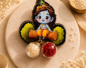 May include: A decorative plaque featuring a cartoon illustration of a blue-skinned child with a peacock feather in their hair, sitting on a brown log. Two small, clear spherical containers hang from the bottom, one filled with white rice and one with red powder.
