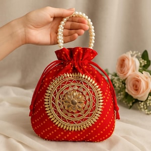 Peut inclure: Un sac rouge à cordon avec une poignée perlée. Le sac présente un motif floral brodé doré et un motif à pois. Le sac est tenu par une main, avec un fond doux et des fleurs.