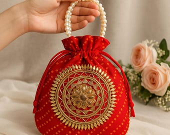 May include: A red drawstring bag with a pearl beaded handle. The bag features a gold embroidered floral design on the front and a dotted pattern. The bag is held by a hand, with a soft background and flowers.
