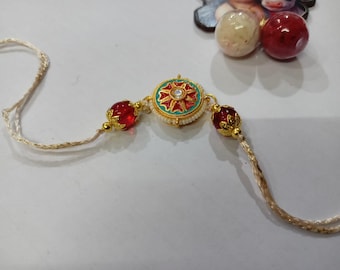 May include: A gold-toned bracelet with a central circular pendant featuring a red and green floral design and a central clear gem. Two red, gem-like beads with gold accents flank the pendant. The bracelet is strung on a cream-colored cord.