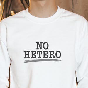 May include: White long-sleeved sweatshirt featuring the text "NO HETERO" in black, underlined with a grey line. The sweatshirt has a crew neck. The person is wearing dark jeans.