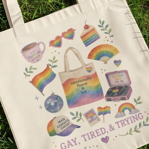 May include: Off-white tote bag with a colorful rainbow design and the text "GAY, TIRED, & TRYING." The bag features various illustrations, including a coffee mug, rainbow flag, and a record player. The bag is sitting on green grass.