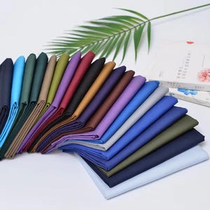 May include: A collection of folded fabric swatches in a wide spectrum of colors including navy, blue, teal, green, tan, olive, purple, maroon, black, brown, and light blue, arranged in a fan-like display on a plain white surface.