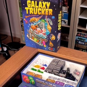 May include: A board game box titled "Galaxy Trucker" with a colorful illustration of a spaceship. The box is open, revealing game components like cards, tokens, and ship tiles. The game is set on a blue surface.