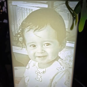 May include: A lit-up framed portrait of a smiling baby. The black-framed picture shows a grayscale image of a child in a light-coloured onesie with a cartoon design. The image is illuminated from within, creating a soft glow.