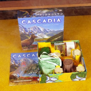 Może przedstawiać: Zestaw gier planszowych "Cascadia" z ilustracją gór i łosia. Otwarta skrzynka ujawnia elementy gry, w tym sześciokątne płytki, drewniane żetony i zielony worek ze sznurkiem. Widoczna jest również książka z zasadami.