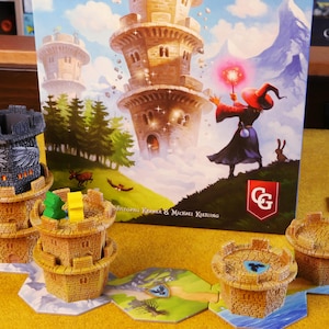 Puede incluir: Un juego de mesa con temática de fantasía. El juego presenta torres de castillo en miniatura en tonos beige y gris, junto con fichas de juego hexagonales. La ilustración de la caja muestra un mago con una varita brillante y un castillo alto.