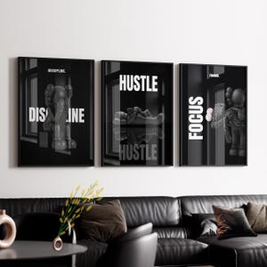 Op de afbeelding: Set van drie zwart-wit ingelijste kunstprints. Elke print toont een KAWS-achtige figuur en motiverende woorden: "DISCIPLINE", "HUSTLE" en "FOCUS". De prints worden weergegeven boven een zwarte leren bank.
