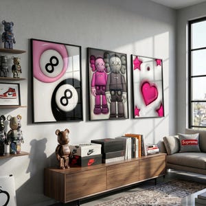 May include: Three framed art prints with black frames. One features a pink and black 8-ball design, another depicts two cartoon figures, and the third showcases a pink heart and stars. A wooden cabinet holds shoe boxes and books.