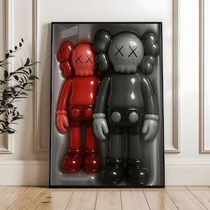 May include: Framed artwork featuring two glossy, stylized figures. One is red, the other is gray, both with distinctive skull-like faces marked with 'XX' eyes. The figures are in a molded plastic display case, set against a white wall.