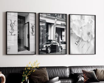 May include: A set of three black and white framed wall art prints. The left print shows a storefront with YSL logos. The center print features a black sports car parked in front of a Chanel boutique. The right print displays white orchids over a Dior book.