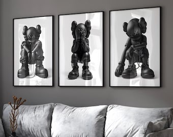 May include: A set of three framed wall art prints featuring black cartoon-style figures with X-shaped eyes. Each print displays a different pose of the same character, including sitting with head down, covering the face, and leaning forward.