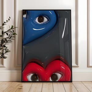 May include: Framed artwork featuring two stylized heart-shaped faces with eyes. The top face is blue, and the bottom face is red, both against a gray background. The artwork is displayed on a wooden floor.