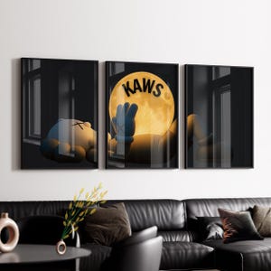 May include: A triptych of framed art prints featuring a cartoon character in a dark setting. One print shows the character lying down, another shows the character holding a glowing moon with the word "KAWS", and the third shows the character's back.
