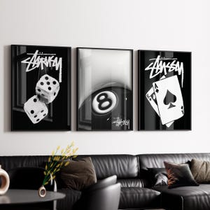 Puede incluir: Un conjunto de tres láminas decorativas enmarcadas en blanco y negro. La lámina de la izquierda muestra dos dados, la central presenta una bola de billar número ocho y la de la derecha muestra dos cartas, un as y un rey de picas. Cada lámina incluye el logo de Stussy.