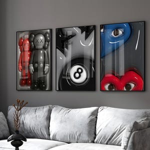 May include: Three framed art pieces with pop art themes. One features a red and gray figure, another a black and white eight ball, and the third a blue eye and red lips. The artwork is displayed above a gray sofa.