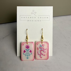 May include: A pair of rectangular drop earrings featuring a glossy finish and gold-tone hooks. One earring displays a pink floral design with the number 3, while the other shows a pink dragon illustration with the number 9, mounted on a card.