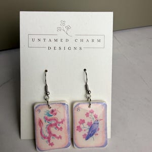May include: A pair of rectangular drop earrings featuring a pink dragon on the left and a purple bird with pink flowers on the right, both labeled with numbers 9 and 8. The earrings are displayed on a white card printed with Untamed Charm Designs.