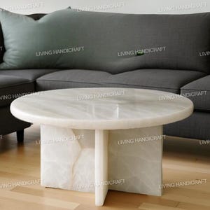 Puede incluir: Una mesa de centro redonda de mármol blanco con una base de pedestal única. La mesa tiene aproximadamente 91 cm de diámetro y mide unos 46 cm de altura. El mármol tiene vetas sutiles. La mesa está frente a un sofá seccional gris.