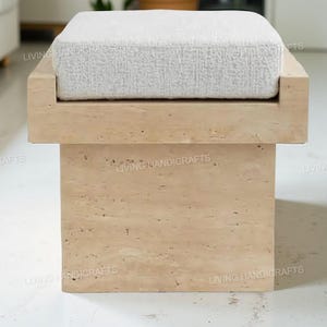 Puede incluir: Un otomán rectangular moderno con una base de piedra beige claro y un cojín blanco texturizado. El otomán tiene un diseño minimalista, adecuado para salones o dormitorios. La base está hecha de piedra de color claro.