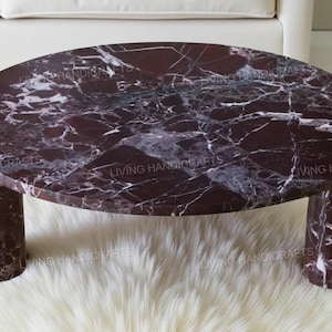 May include: Oval-shaped coffee table with a dark burgundy marble top and cylindrical legs. The marble features white veining, creating a striking contrast. The table is set on a white, fluffy rug.