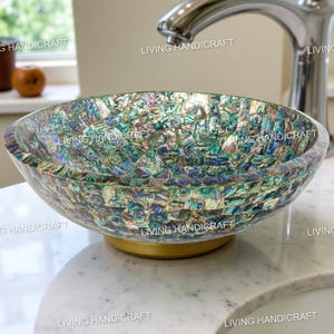 May include: A decorative, oval-shaped bathroom sink with a mosaic pattern of iridescent blue, green, and gold pieces. The sink sits on a gold-colored base. A chrome faucet is visible in the background. The text "LIVING HANDICRAFT" is visible on the sink.