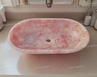 以下が含まれることがあります： ピンクと白の石で作られた楕円形の洗面台。洗面台は、中央に排水穴があり、滑らかで研磨された表面をしています。洗面台は明るい色のカウンタートップに置かれています。「LIVING HANDICRAFT」の文字が洗面台に印刷されています。