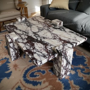 Puede incluir: Una mesa de centro rectangular de mármol blanco con vetas moradas oscuras. La mesa tiene un diseño único con patas y soportes geométricos. La mesa de mármol es una pieza destacada para una sala de estar.