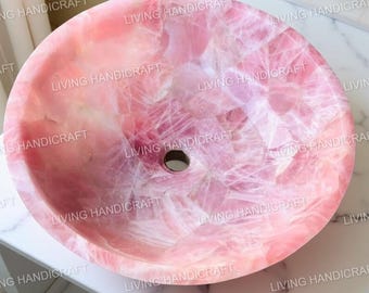 Pode incluir: Uma pia redonda de quartzo rosa com um padrão natural e veteado. Possui um orifício de drenagem central e um acabamento polido. As palavras "LIVING HANDICRAFT" são impressas na borda.