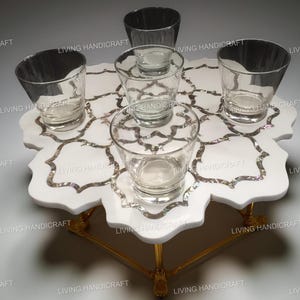 Puede incluir: Una bandeja de mármol blanco con un diseño floral con incrustaciones de detalles plateados. Seis vasos transparentes están dispuestos en la bandeja, que está sostenida por patas doradas. Se ven las palabras "LIVING HANDICRAFT".