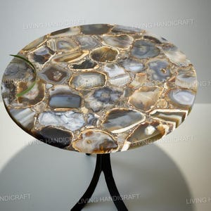 May include: A round side table with a natural stone top. The tabletop is a mosaic of beige, grey, and brown stones. The table has a black metal base with three curved legs. A green plant is on the table.