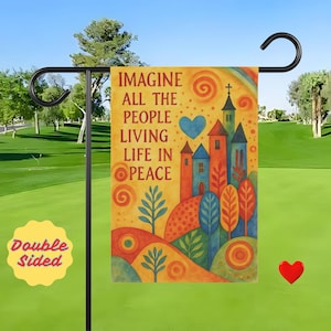 May include: A double-sided garden flag with the text "IMAGINE ALL THE PEOPLE LIVING LIFE IN PEACE" on a yellow background. The flag features a colorful illustration of a town with a rainbow, trees, and a blue heart. The flag is hanging on a black metal stand.