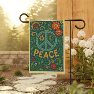 May include: A garden flag with a blue background features a peace symbol and the word "PEACE" in yellow. The flag is decorated with colorful flowers and green foliage. The flag is displayed on a black metal stand in a garden setting.