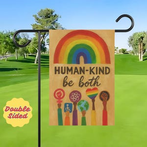 May include: A double-sided garden flag with a rainbow and the words "HUMAN-KIND be both." The flag features various symbols of inclusivity, including a fist, a heart, and a globe. The flag is displayed on a black metal stand.