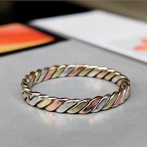 May include: A multi-tone ring with a twisted design. The ring features alternating sections of gold, rose gold, and silver metal. The ring is set against a light gray surface.