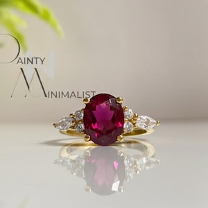 May include: A gold ring with a large oval ruby centre stone, and smaller pear-shaped diamonds. The ring has a delicate band. The ring is a piece of jewellery. The background is soft and neutral.