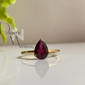 Op de afbeelding: Een gouden ring met een peervormige, dieprode edelsteen. De ring heeft een dunne gouden band en de edelsteen wordt op zijn plaats gehouden door een kleine gouden poot. De ring wordt getoond op een wit oppervlak met een zachte reflectie.