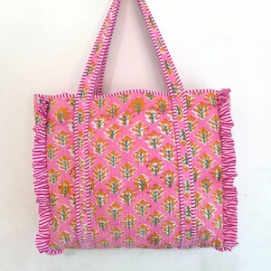 May include: Pink tote bag with a floral pattern in orange and white. The bag has matching pink handles and ruffled edges. The bag is made of a quilted fabric.
